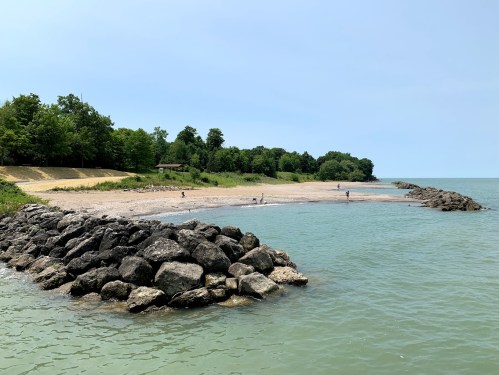 A rocky shoreline extends into calm waters, with a sandy beach and trees in the background under a clear sky.
