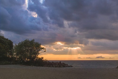 A serene beach scene at sunset, with dark clouds and rays of light breaking through over the calm ocean.