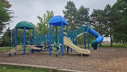 A colorful playground with slides and climbing structures, surrounded by trees and a grassy area on a cloudy day.