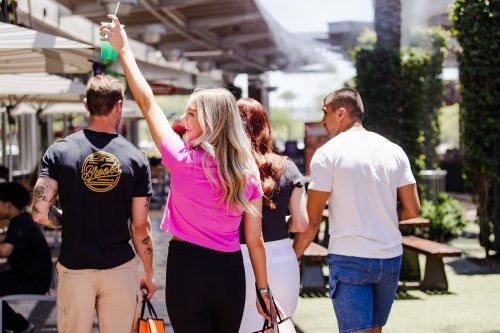 A group of four people walking outdoors, one woman in a pink shirt raises a drink, while others carry shopping bags.