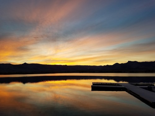A serene lake at sunset, with vibrant colors reflecting on the water and a silhouette of mountains in the background.