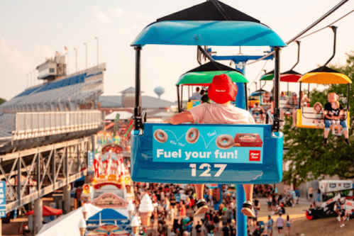 A person in a red hat rides a blue chairlift above a bustling fairground filled with colorful attractions.