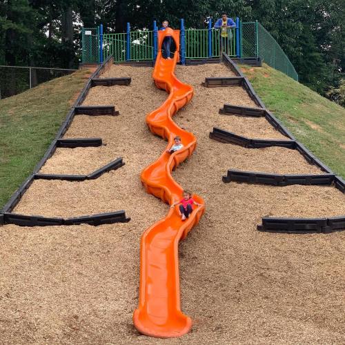 Two orange slides on a grassy hill at a playground, with children playing and climbing.