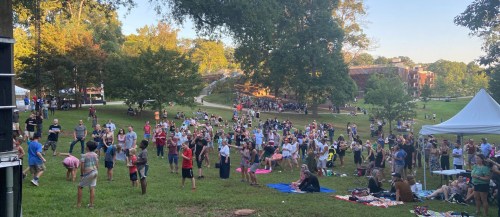 A large crowd enjoys an outdoor event in a park, with people gathered on the grass and under trees.