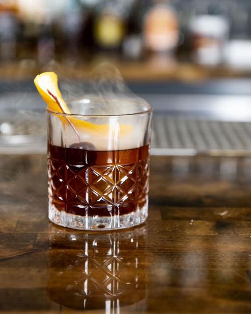 A cocktail in a crystal glass, garnished with an orange slice and cherry, sits on a wooden bar surface.
