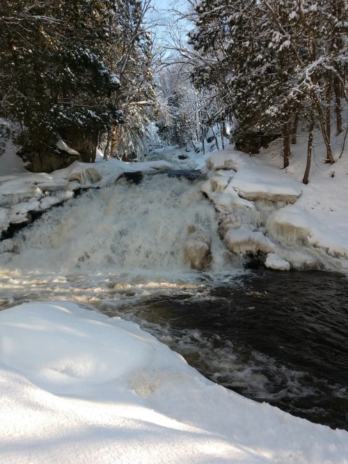 A snowy landscape featuring a waterfall cascading over ice and rocks, surrounded by trees.