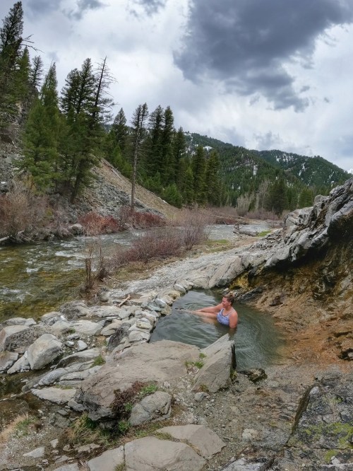 A person relaxes in a natural hot spring surrounded by rocky terrain and lush green trees under a cloudy sky.