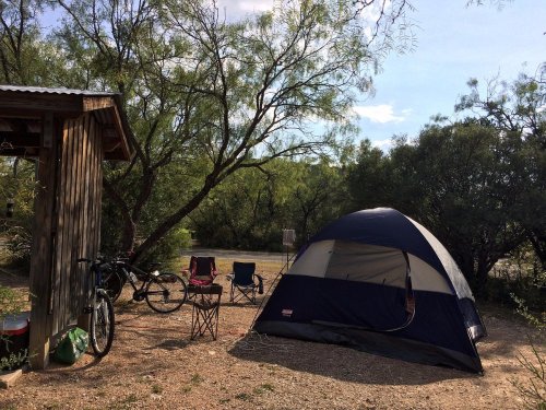 A blue tent set up in a campsite surrounded by trees, with chairs and a bicycle nearby.