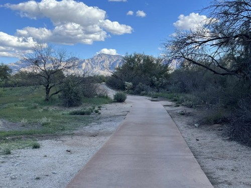 A winding path through a green landscape, with mountains and clouds in the background under a blue sky.
