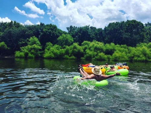 People floating on colorful inner tubes in a river, surrounded by lush greenery and a bright blue sky.