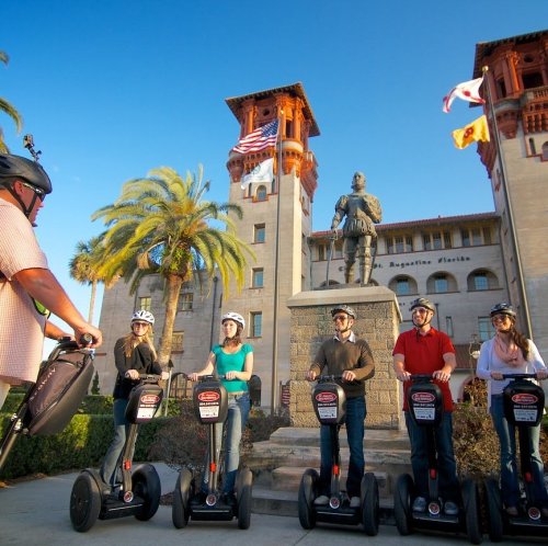 A group of people on Segways in front of a historic building and statue, with palm trees and flags in the background.