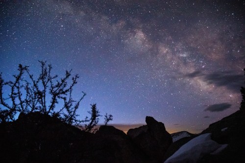 A starry night sky with the Milky Way visible, framed by silhouetted rocks and sparse vegetation.