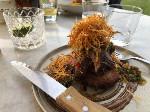A plate of steak topped with crispy fried onions and sauce, served with a knife and fork on a marble table.