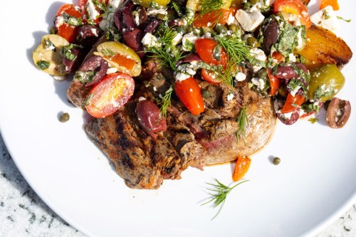 A plate of grilled meat topped with colorful tomatoes, olives, feta cheese, and fresh herbs.