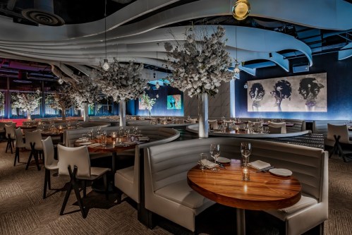 Modern restaurant interior with elegant seating, floral arrangements, and artistic wall decor. Soft lighting creates a cozy atmosphere.