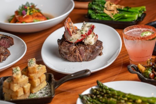 A gourmet meal featuring a steak topped with lobster, surrounded by sides of asparagus, potatoes, and a cocktail.