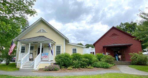A yellow house with an "Open" sign and a red building nearby, surrounded by greenery under a cloudy sky.