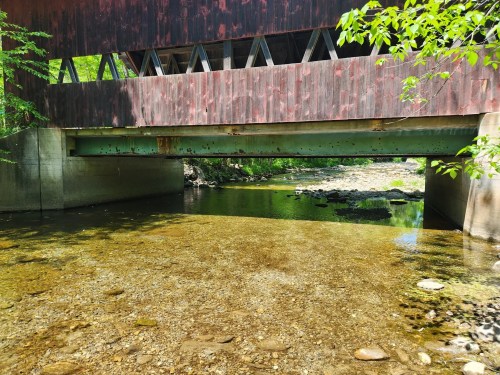 A covered bridge spans a shallow, clear stream with rocky banks and greenery surrounding the scene.