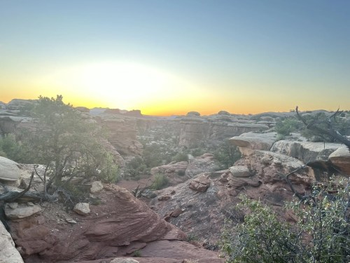 Sunset over a rocky landscape, with vibrant orange and yellow hues illuminating the sky and rugged terrain.