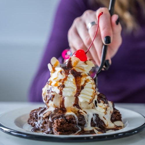 A hand holds a fork above a dessert with brownie, ice cream, whipped cream, chocolate sauce, and a cherry on top.
