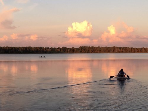 A person rows a small boat on a calm lake at sunset, with soft clouds reflecting in the water.