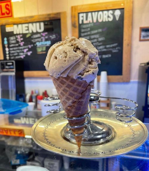 A cone of ice cream sits on a silver platter, with a chalkboard menu in the background displaying various flavors.