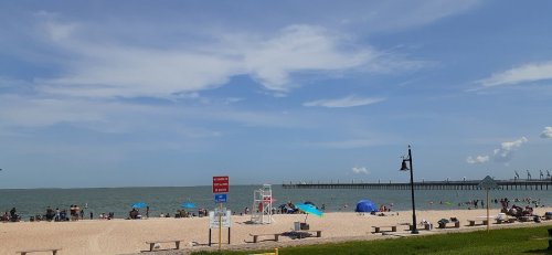 A beach scene with people relaxing on the sand, colorful umbrellas, and a pier in the background under a blue sky.