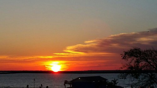 A vibrant sunset over a calm sea, with orange and yellow hues illuminating the sky and silhouetting a small building.