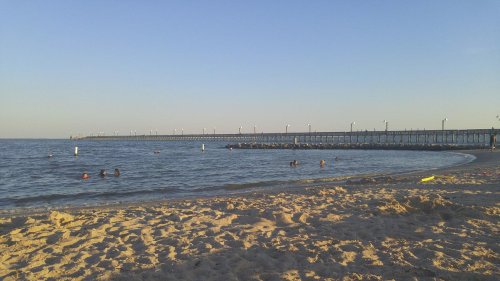 A sandy beach with people swimming in the water and a long pier extending into the sea under a clear blue sky.