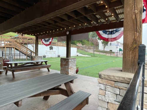 A covered outdoor area with picnic tables, string lights, and patriotic decorations, overlooking a grassy field.