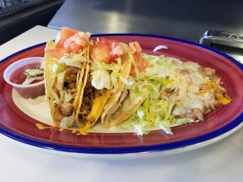 A plate of two tacos filled with meat, cheese, and lettuce, served with a side of refried beans and a small cup of salsa.