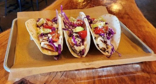 Three fish tacos on a tray, topped with cabbage, tomatoes, and cheese, served on a wooden table.