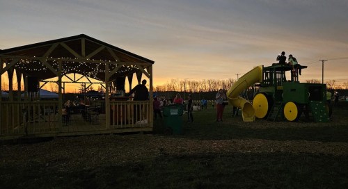 A playground with a tractor slide and a gazebo, set against a colorful sunset. People are enjoying the outdoor space.