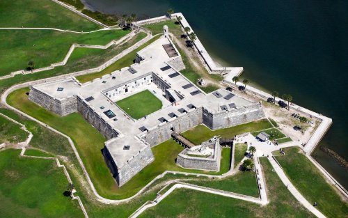 Aerial view of a historic fort surrounded by green lawns and water, showcasing its star-shaped design and stone walls.