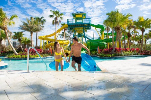 A couple in swimwear joyfully enters a pool, holding inflatable tubes, with a colorful water slide and palm trees in the background.