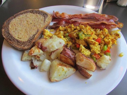 A plate of breakfast featuring scrambled eggs with peppers, bacon, roasted potatoes, and a slice of whole grain bread.