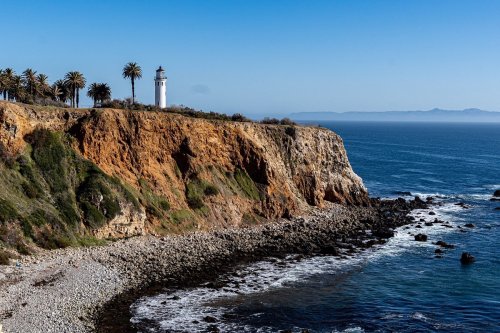 A lighthouse on a rocky cliff overlooking the ocean, surrounded by palm trees and clear blue skies.