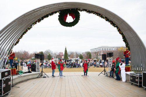 A festive outdoor event with performers on stage, surrounded by a crowd and holiday decorations, including a large wreath.