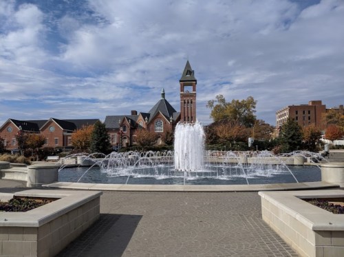 A fountain sprays water in a plaza, with a historic building and trees in the background under a cloudy sky.