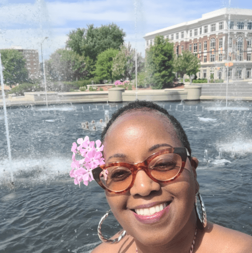 A smiling woman with glasses and a pink flower in her hair stands by a fountain on a sunny day.