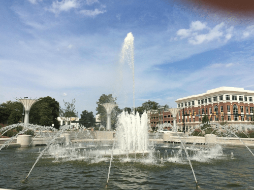 A fountain sprays water into the air, surrounded by greenery and buildings under a blue sky with scattered clouds.