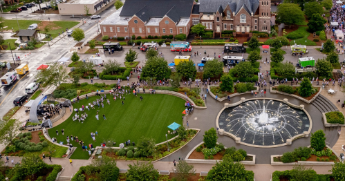 Aerial view of a park with a fountain, food trucks, and people gathered on the lawn in a vibrant urban setting.
