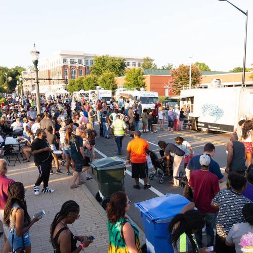 A large crowd gathers at a street event with food trucks, enjoying the festivities on a sunny day.