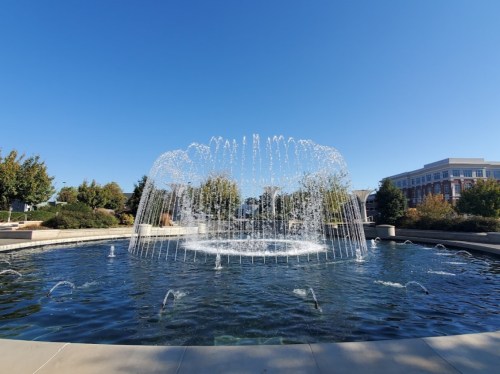 A large fountain sprays water in a circular pattern, surrounded by greenery and a clear blue sky.