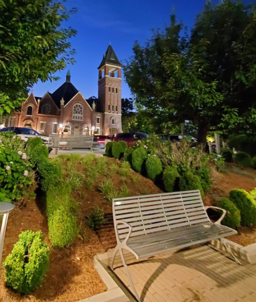A park bench in front of a church at dusk, surrounded by neatly trimmed bushes and trees.