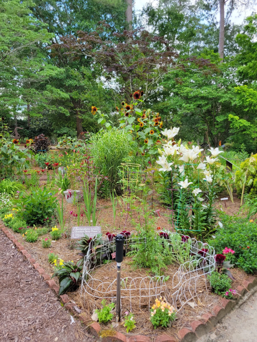 A vibrant garden filled with various flowers, including sunflowers and lilies, surrounded by greenery and decorative fencing.