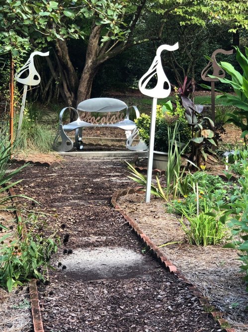 A garden path leads to a silver bench surrounded by decorative sculptures and lush greenery.