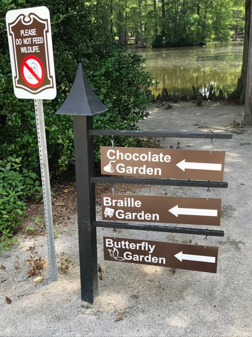 Signpost with directions to Chocolate Garden, Braille Garden, and Butterfly Garden near a pond, with a wildlife warning.