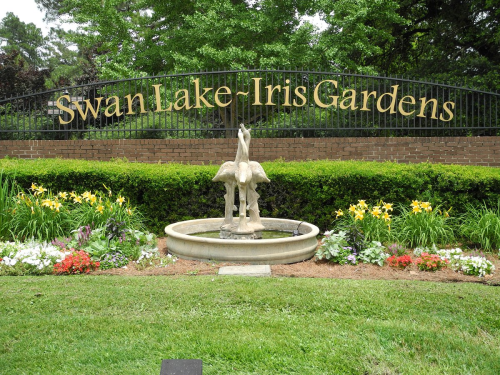 Sign for Swan Lake Iris Gardens with a fountain and colorful flowers in front. Lush greenery surrounds the area.