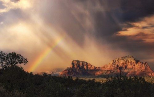 A vibrant rainbow arcs over red rock formations under a dramatic sky with rays of sunlight breaking through clouds.